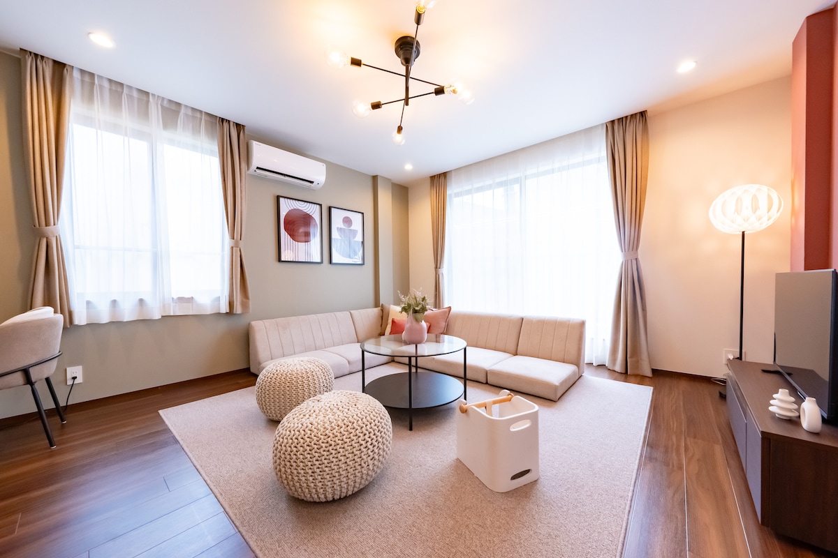 A spacious living area features large windows with sheer curtains, allowing natural light to brighten the room. A round coffee table sits on a soft area rug, surrounded by a sectional sofa and two textured poufs, creating a comfortable gathering space.