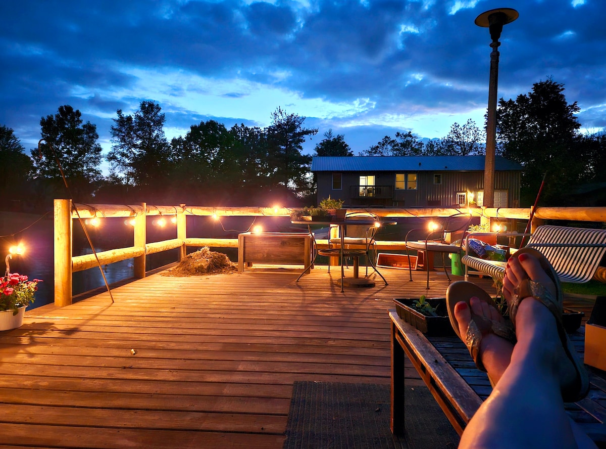 A wooden deck is illuminated by string lights, creating a warm atmosphere in the evening. Comfortable seating is arranged around a table, with distant views of trees and a building across the pond. A comfortable sitting area with feet resting on a nearby bench is visible.