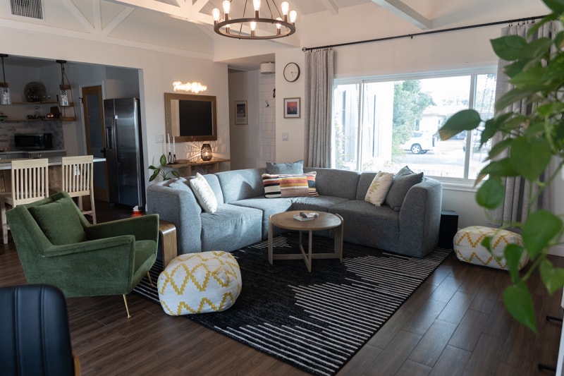 A spacious common area showcases a large, gray sectional sofa arranged around a round coffee table. Two armchairs in contrasting green and patterned poufs enhance the seating options. A large window invites natural light, while decorative touches, such as plants and wall art, add character.