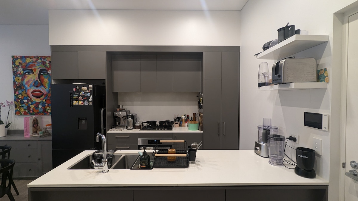 The modern kitchen features sleek cabinetry and a spacious island with a sink. Cooking appliances, including a stovetop and oven, are visible. Multiple countertop gadgets, such as blenders and a coffee machine, are arranged neatly, contributing to a functional cooking space.