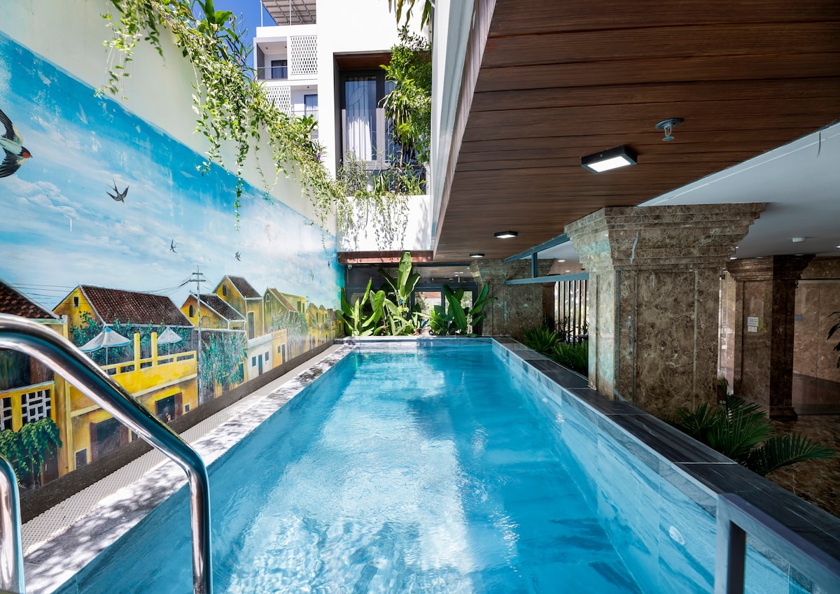 An indoor swimming pool is surrounded by lush greenery and vibrant murals depicting local architecture. The pool area features smooth stone tiles and elegant lighting, creating a serene environment for relaxation and unwinding.