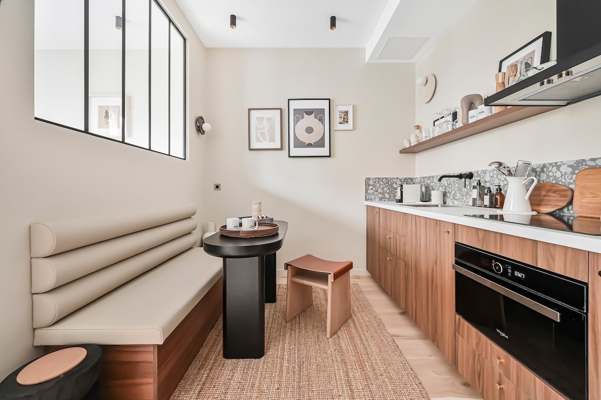 A modern kitchen area features a wooden countertop and an integrated oven. A dining space is provided with a round black table and a curved bench, complemented by an accent stool. The walls are adorned with framed artwork, adding a touch of style.