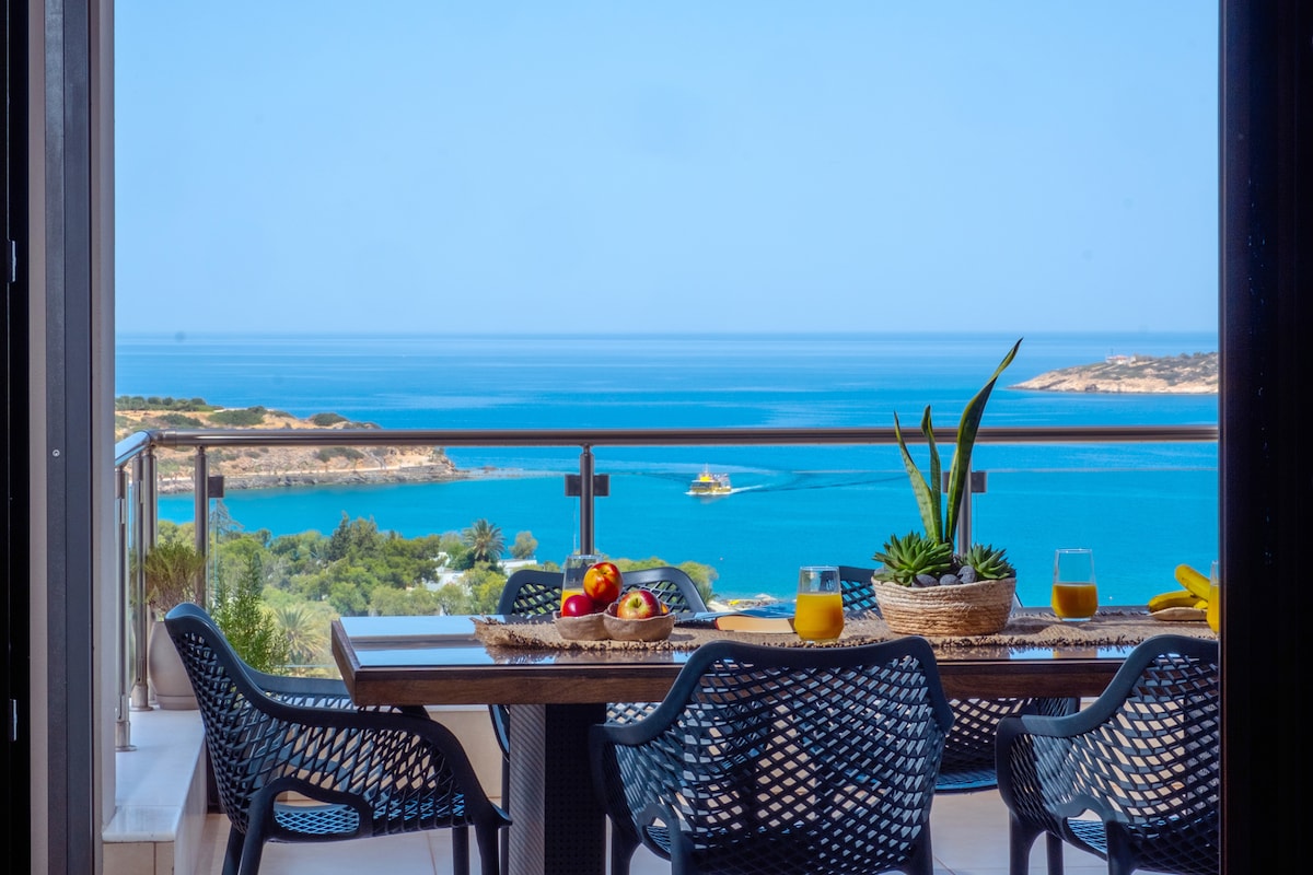 A dining table is positioned on a balcony, offering panoramic views of the sea. Four woven chairs surround the table, which is adorned with fresh fruit, a jug, and glasses. The serene blue waters and distant land add to the scene's tranquility.
