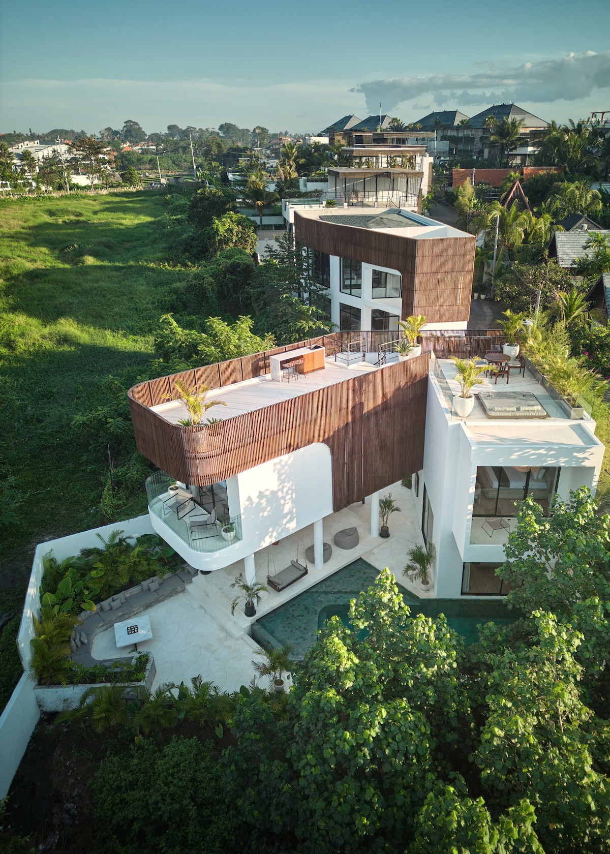 An aerial view of the luxury villa showcases its modern architecture with distinct, layered structures. The villa is surrounded by lush greenery, providing a serene backdrop. A rooftop area can be seen, complemented by outdoor seating, while the private pool adds to the inviting atmosphere of the property.