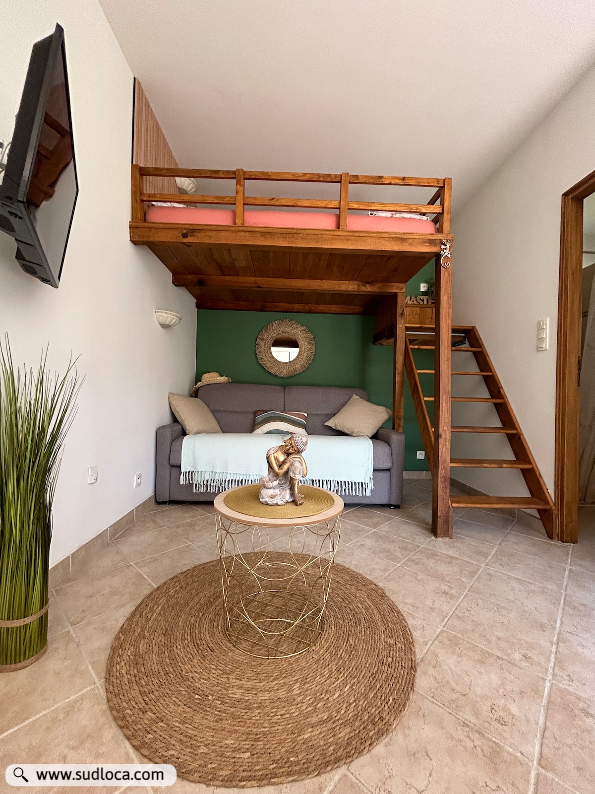A cozy studio space is featured, showcasing a lofted sleeping area above a comfortable sofa. A decorative plant and textured area rug add warmth to the tile floor. A small, round table occupies the center, enhancing the welcoming atmosphere of the room.