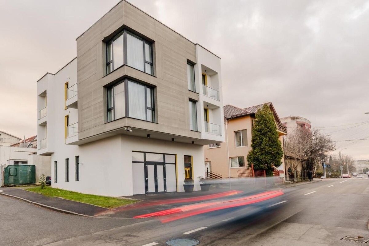 A modern three-story building features a sleek design with large windows and a neutral facade. The structure is situated at a street corner, offering access points along the sidewalk. Nearby, typical residential architecture contrasts with the contemporary style.