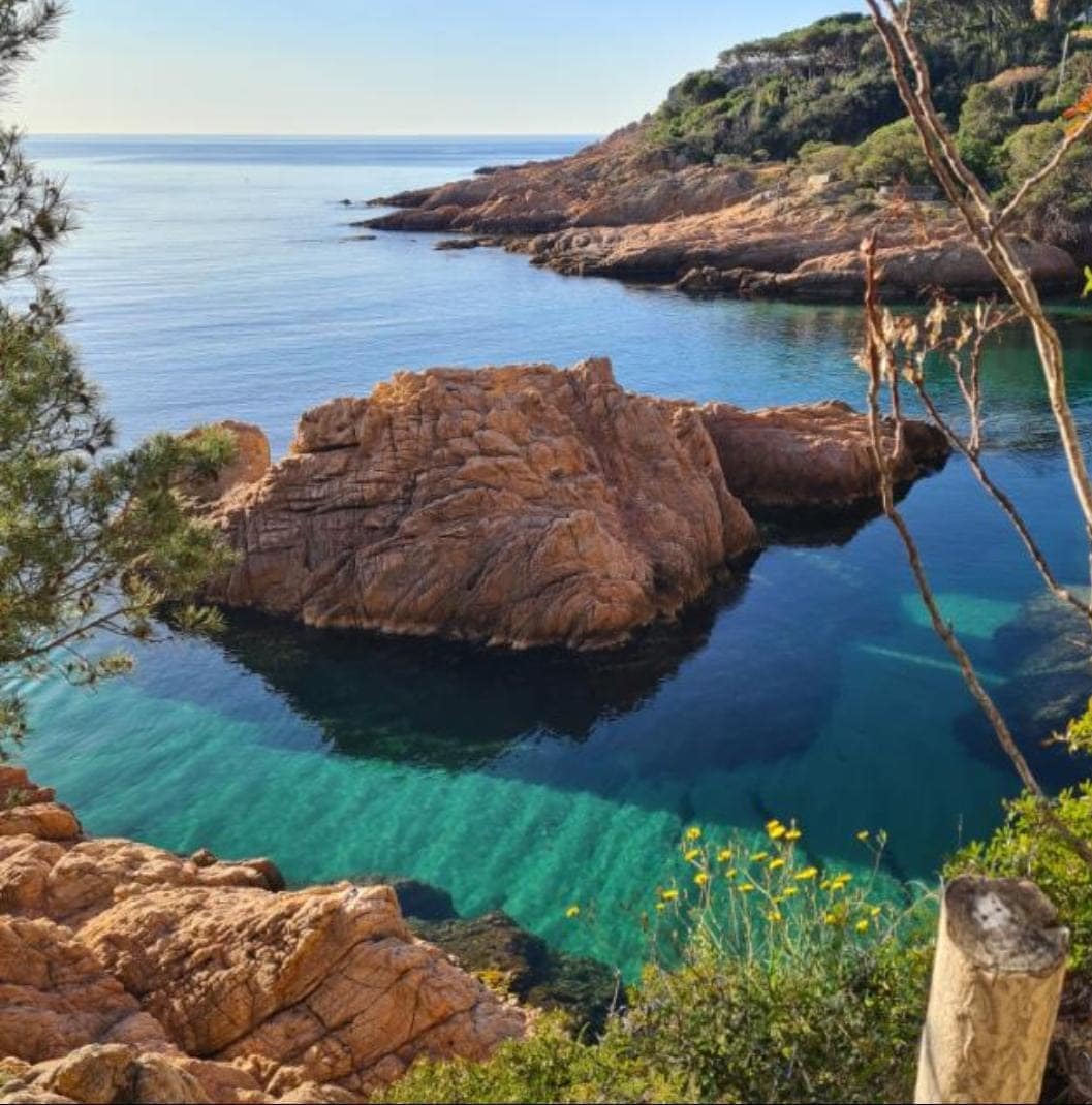 A rocky coastline features clear turquoise water gently lapping against the shore. A large rock formation juts out from the water, surrounded by a natural landscape of greenery. The tranquility of the scene is enhanced by the calm surface reflecting the coastal surroundings.