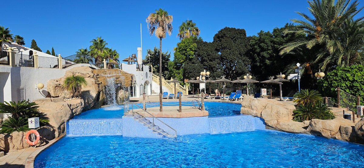 A multi-level pool area features cascading waterfalls amidst landscaped surroundings including palm trees. Several sun loungers are positioned nearby for relaxation, with an inviting blue water reflecting the clear sky. The area is well-lit with wall lights for an enjoyable atmosphere during the evening.