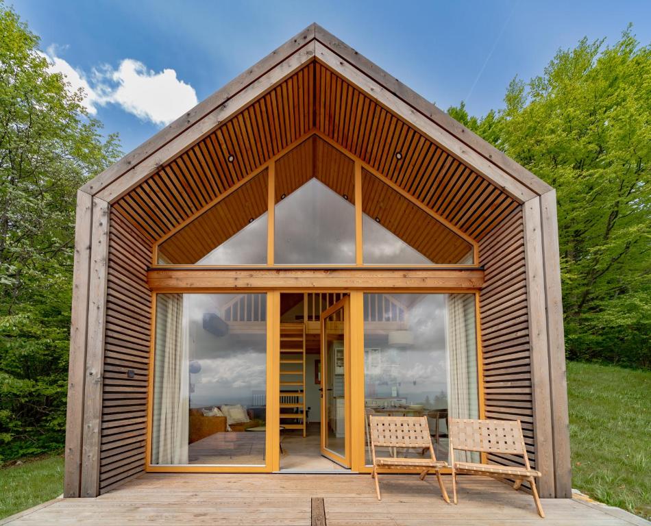 A modern wooden cabin features a steeply pitched roof with large glass doors that open to a spacious deck. Two wooden chairs are placed on the deck, providing a relaxing spot to enjoy the views of the surrounding greenery.