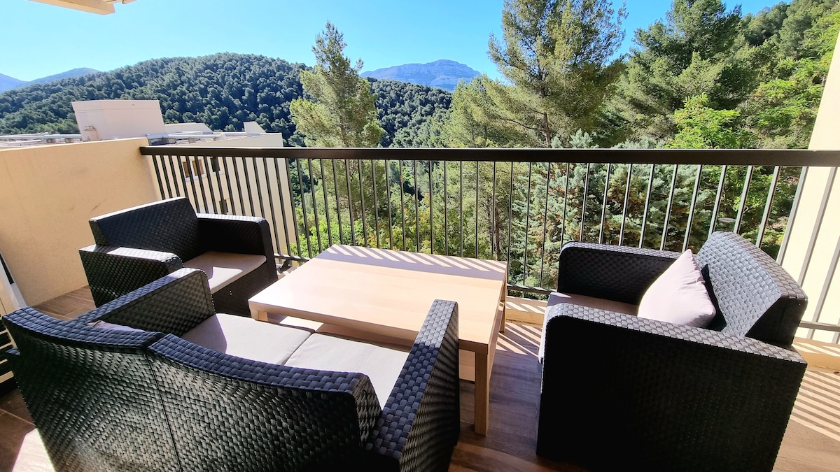A furnished terrace features a table surrounded by four wicker chairs, offering a view of the surrounding greenery and mountains. The railing provides a sense of openness while maintaining privacy.