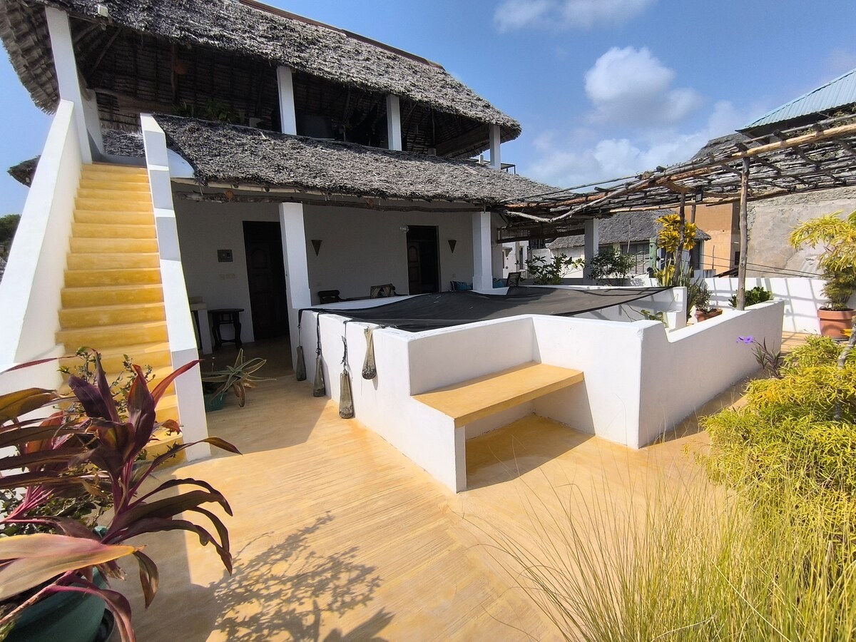 The outdoor terrace is framed by lush greenery, featuring a wooden bench and decorative plants. A staircase leads to the upper levels of the townhouse, while traditional thatched roofing adds a touch of local charm to the structure.