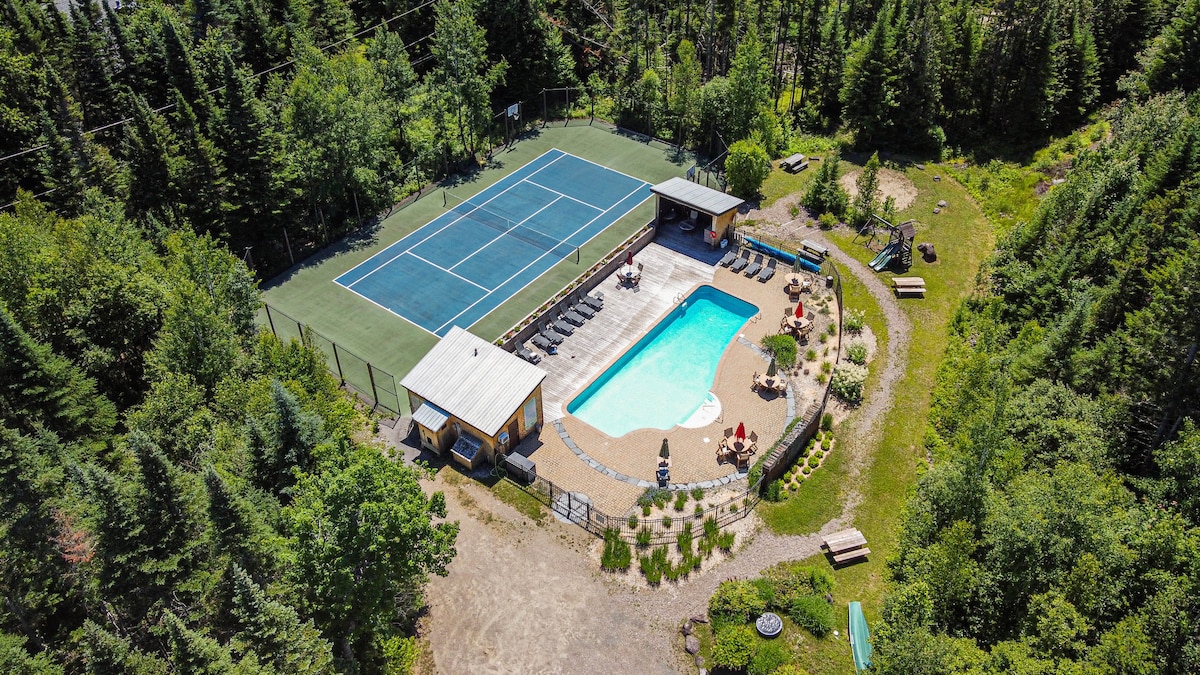 Aerial view of a recreational area surrounded by trees, featuring a tennis court and a swimming pool. Lounge chairs are arranged around the pool, with shaded areas provided by nearby structures. A natural landscape complements the tranquil setting, inviting relaxation and outdoor activities.
