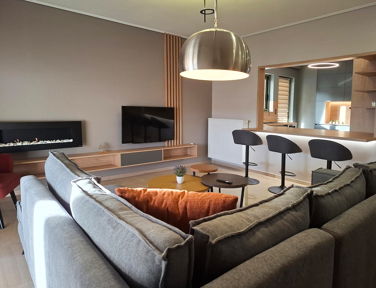 A spacious living area features a sectional sofa accented with decorative pillows and a warm-colored throw. A modern lighting fixture hangs above a set of sleek stools positioned around a circular coffee table. A television is mounted on a wall unit, enhancing the contemporary design.