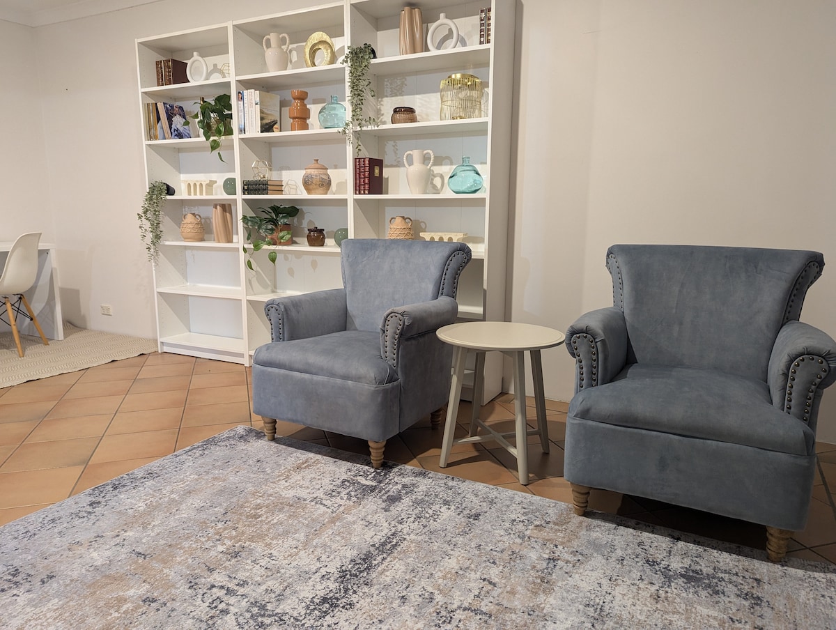 A cozy seating area features two blue upholstered chairs with decorative nailhead trim, positioned alongside a small round table. A neutral-toned area rug covers the tiled floor, while a white bookshelf in the background displays various decorative items and plants.