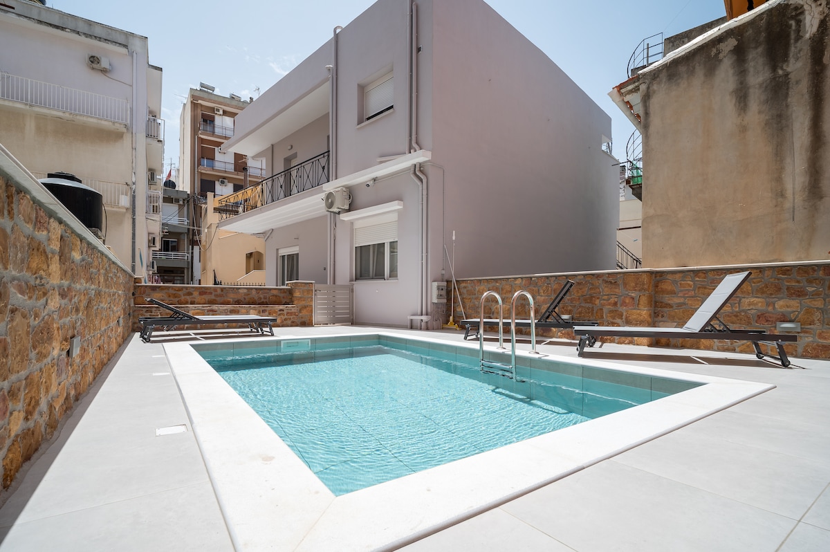A well-maintained outdoor pool is surrounded by a smooth tiled deck. Several lounge chairs are positioned around the pool for relaxation. The setting is framed by nearby buildings, providing a cozy atmosphere for guests to unwind.