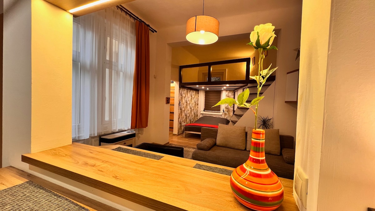 A vibrant flower vase is placed on a wooden countertop, adding a pop of color. The open layout reveals a cozy seating area with a sofa and a glimpse of the sleeping space beyond, illuminated by natural light from large windows adorned with sheer curtains.