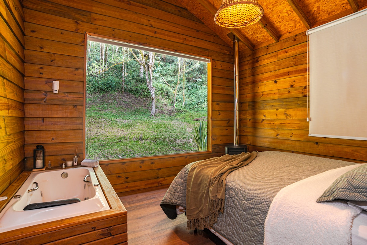 The interior features warm wooden walls and a spacious bed draped in soft linens. A luxurious hot tub sits adjacent to the bed, while a large window offers a view of the lush greenery outside, enhancing the serene atmosphere.