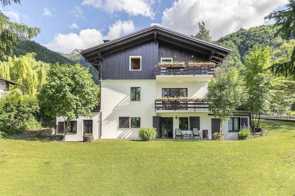 A spacious garden apartment is set against a lush green backdrop, surrounded by rolling hills. Balconies adorned with flowers enhance the property. The outdoor area features a well-maintained lawn with several seating options arranged on a patio, inviting guests to relax in nature.