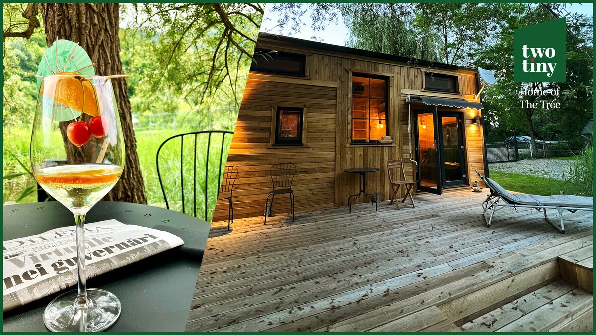 A wooden deck is highlighted, showcasing a cozy seating area with two chairs and a reclining lounge. The cabin's exterior features natural wood siding, with warm lighting creating an inviting look. A cocktail glass sits elegantly on a table, adorned with garnishes and a newspaper.