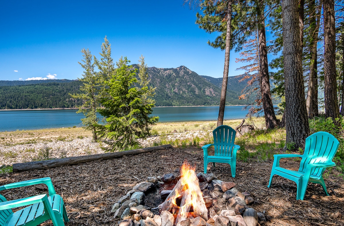 A fire pit surrounded by several teal chairs is positioned near a tranquil lake. Lush trees frame the scene, while distant mountains rise against a clear blue sky. The calm water reflects the natural beauty of the area, inviting relaxation and connection with nature.