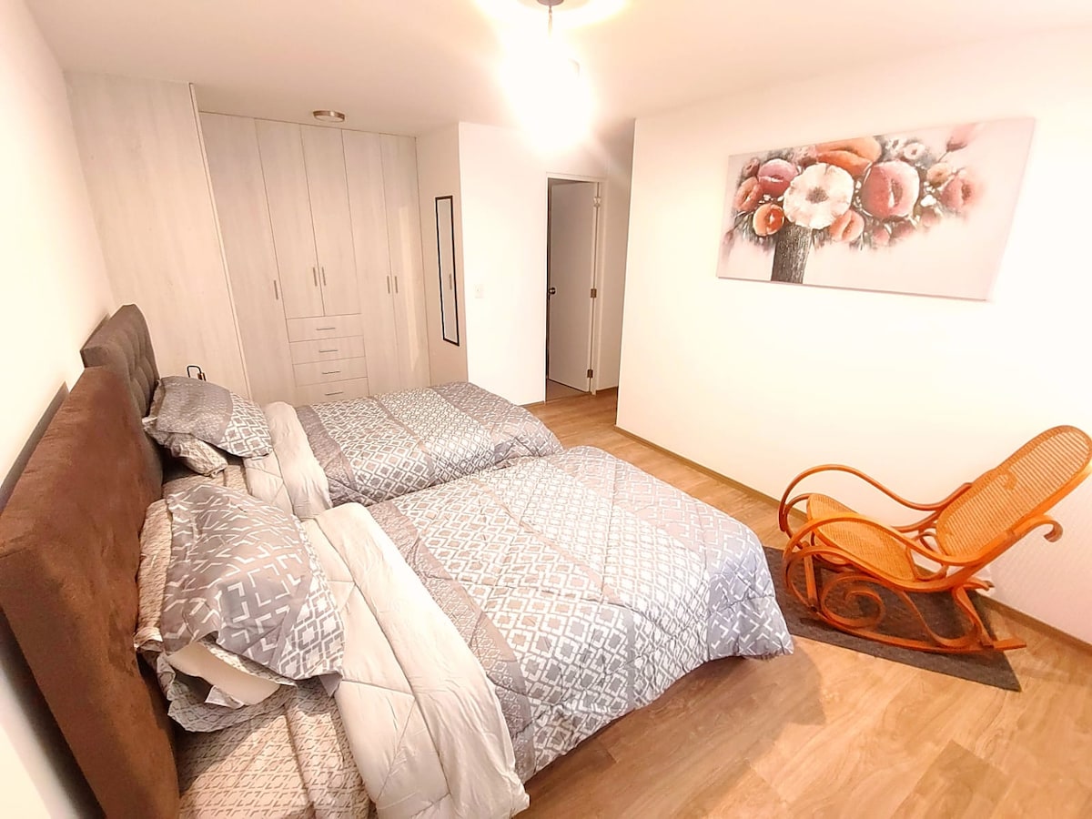 A well-lit bedroom features two separate beds dressed in patterned bedding. A decorative wall art piece enhances the space, while a rocking chair adds a cozy touch. Ample storage is available with built-in wardrobes, creating a tidy and organized environment.