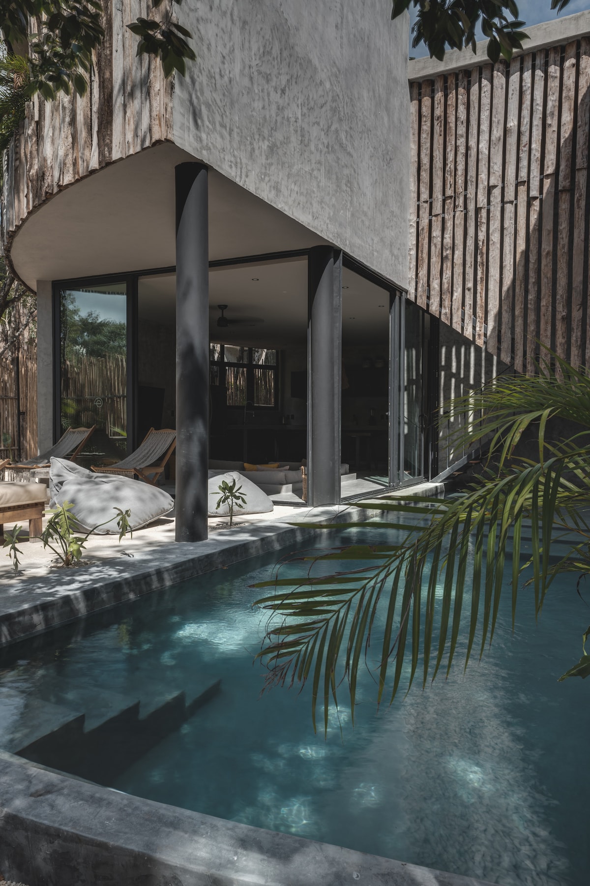 The image showcases a modern villa featuring a private pool surrounded by tropical greenery. Large glass doors create a seamless connection between indoor and outdoor spaces. Textured wooden walls add depth to the architecture, while sun loungers provide a space for relaxation.