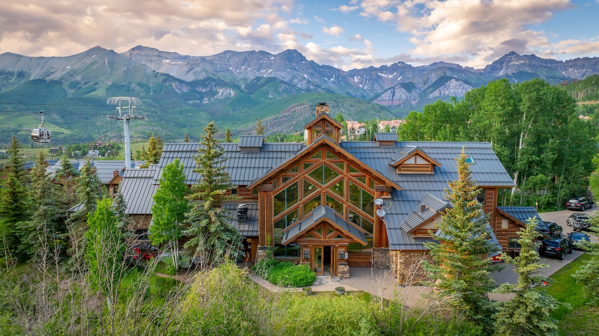 The lodge features a large, glass-fronted design, allowing natural light to fill the interior. Surrounded by lush greenery, the building is set against a backdrop of mountains. Nearby, a ski lift can be seen, indicating proximity to winter activities.