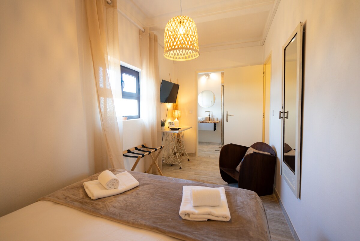 A comfortable suite features a spacious bed layered with soft linens. Natural light filters through sheer curtains, illuminating the room. A TV is mounted on the wall, and a small table with two chairs is positioned nearby. A modern bathroom can be seen through a door.