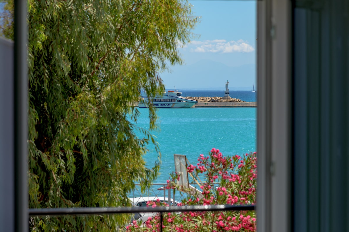 A view from the window showcases a vibrant turquoise sea and a boat anchored near a scenic pier. Lush greenery and blooming flowers frame the scene, adding natural color and tranquility to the coastal landscape.