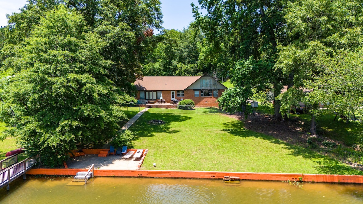 A spacious lakefront property is set against a backdrop of lush greenery. The flat lawn leads to a serene water edge, where a small patio area with seating is visible. The house, with a large deck and multiple windows, offers direct access to the picturesque landscape.