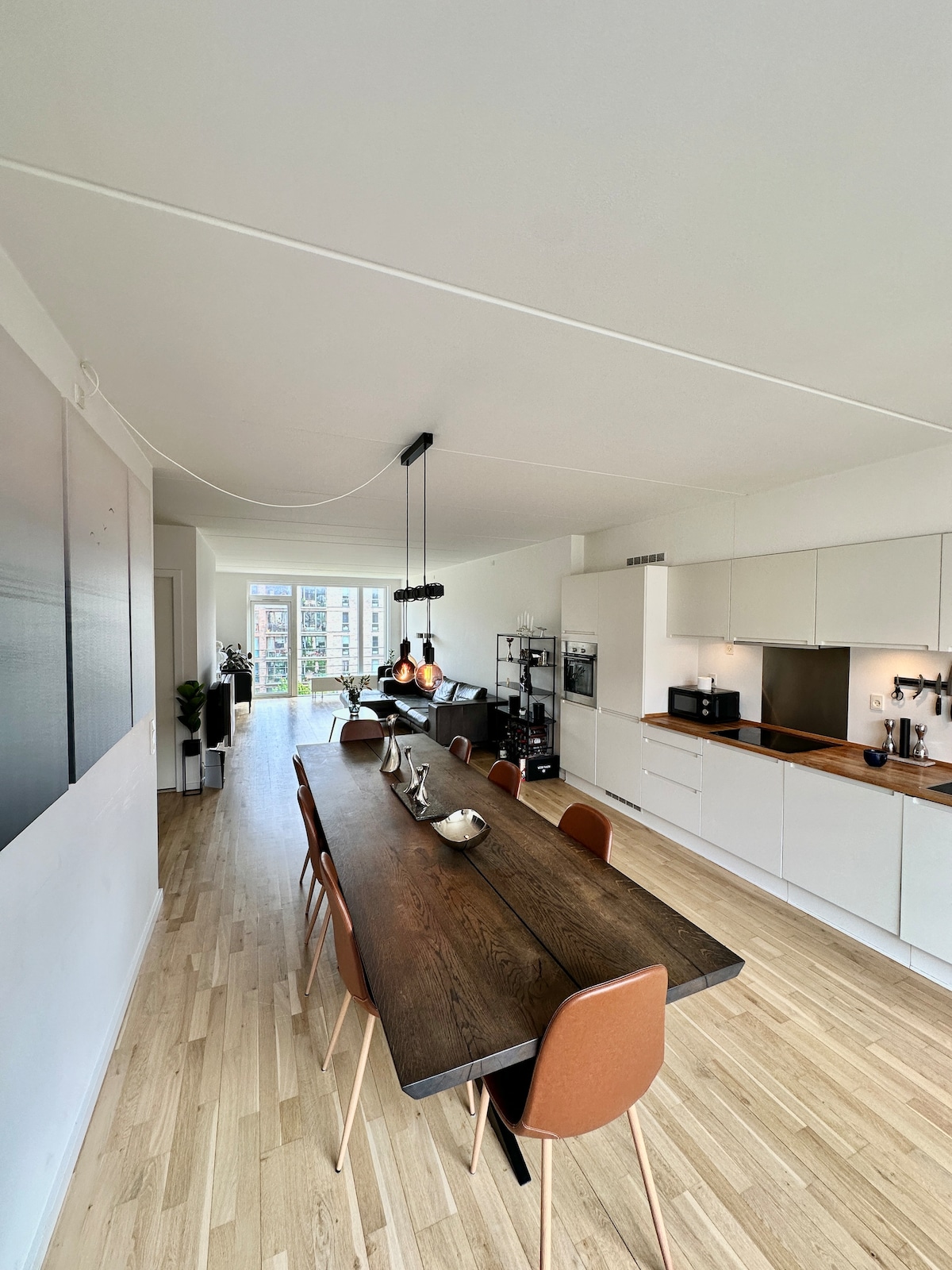 A modern dining area features a long wooden table paired with several brown chairs. Large windows allow natural light to fill the space, illuminating the minimalist kitchen and inviting atmosphere. Contemporary decor lines the walls, enhancing the room's spacious feel.