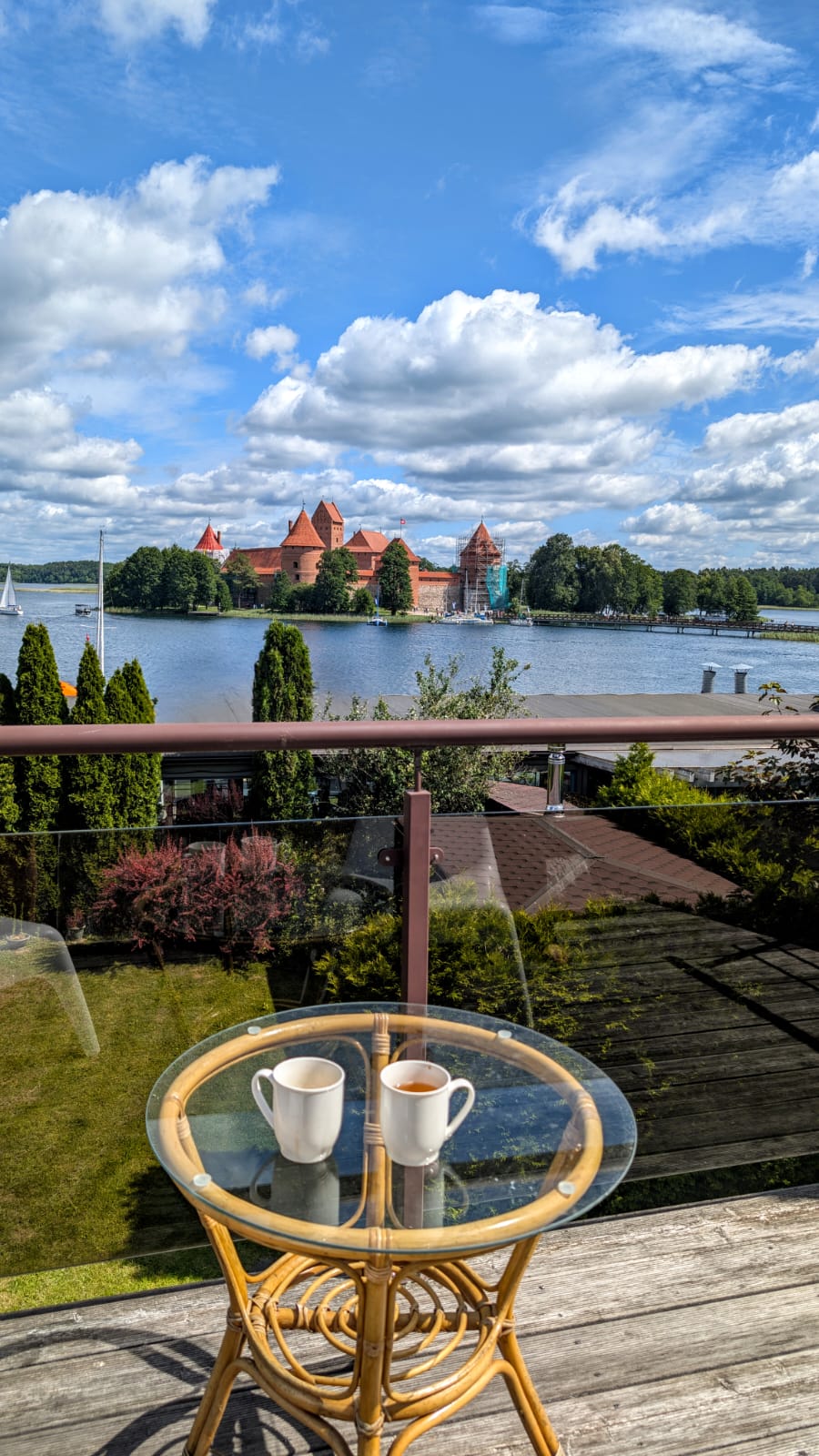 A terrace view shows a charming table with two cups on it, overlooking a lake. The picturesque backdrop features the medieval Trakai Island castle, framed by lush greenery and a bright sky with clouds.