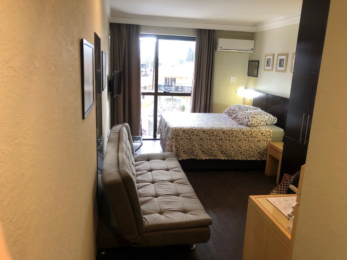 A compact living space features a spacious bed adorned with a floral-patterned coverlet. A seating area includes a gray sofa bed positioned beside a wooden desk. Natural light fills the room through a large window, framed by beige curtains. A wall-mounted television is also visible.