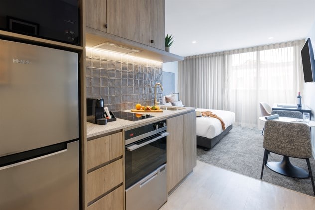 A compact kitchen is featured, equipped with modern appliances including a refrigerator and stovetop. The open layout connects to a sleeping area with a spacious bed. Natural light filters through sheer curtains, illuminating a comfortable seating area with a desk and chair.