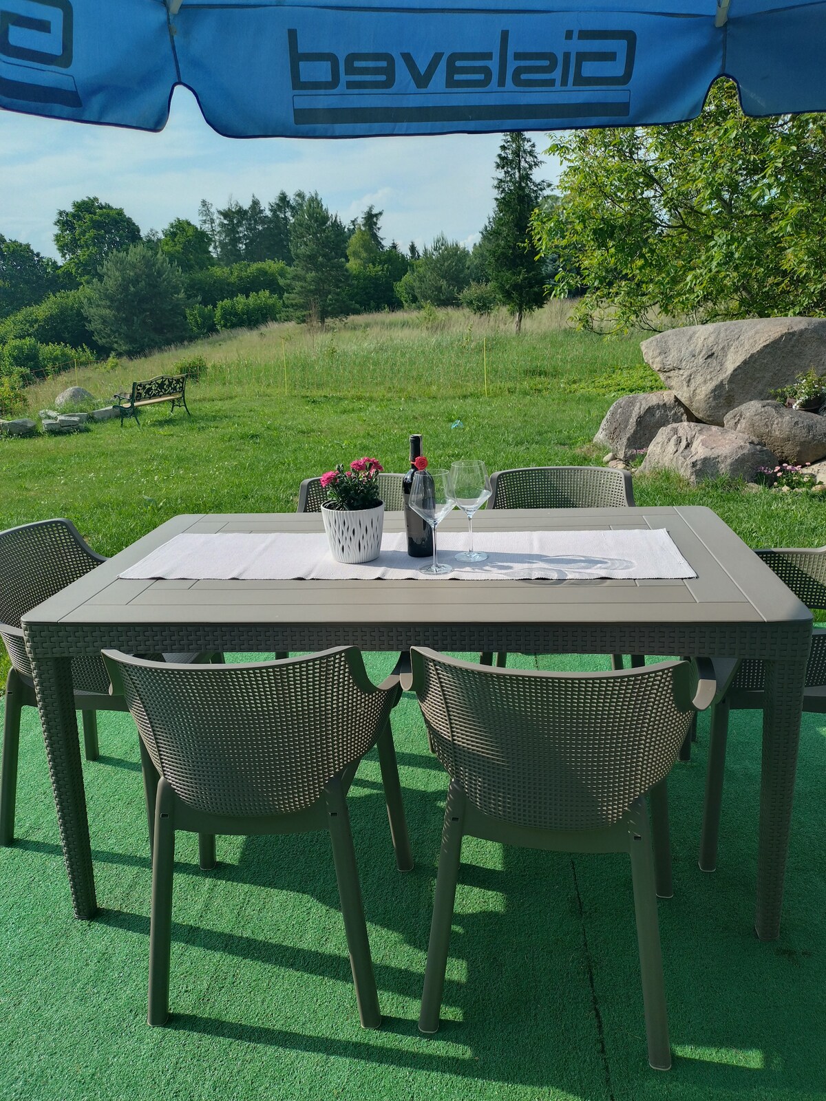 A dining area is set outside under a large umbrella, featuring a gray table with a white table runner. Several chairs surround the table, and a small pot of flowers is placed in the center, with a bottle of wine and two glasses ready for enjoyment. The greenery of the surrounding landscape is visible.