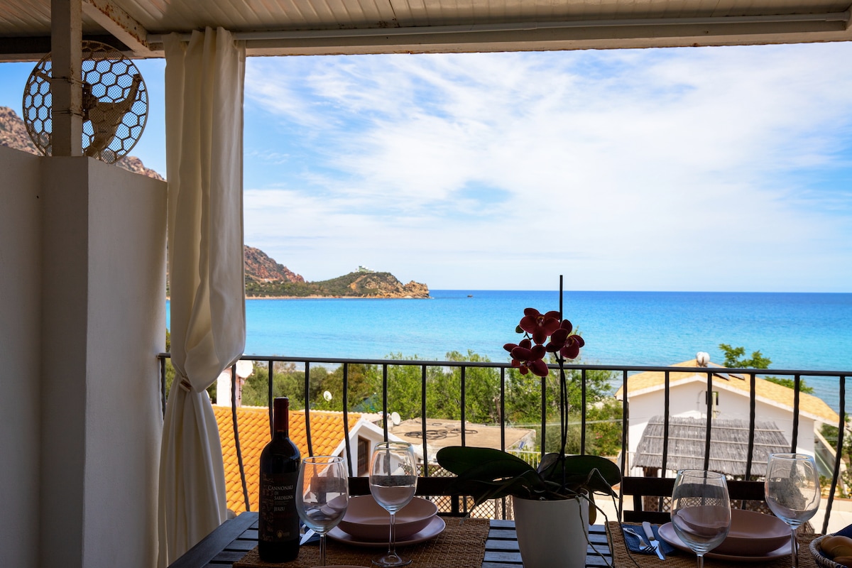 A dining area is visible through a balcony, featuring a table set with wine glasses and a decorative orchid. The expansive sea view showcases a clear blue sky and coastal landscape, enhancing the ambiance for outdoor meals.