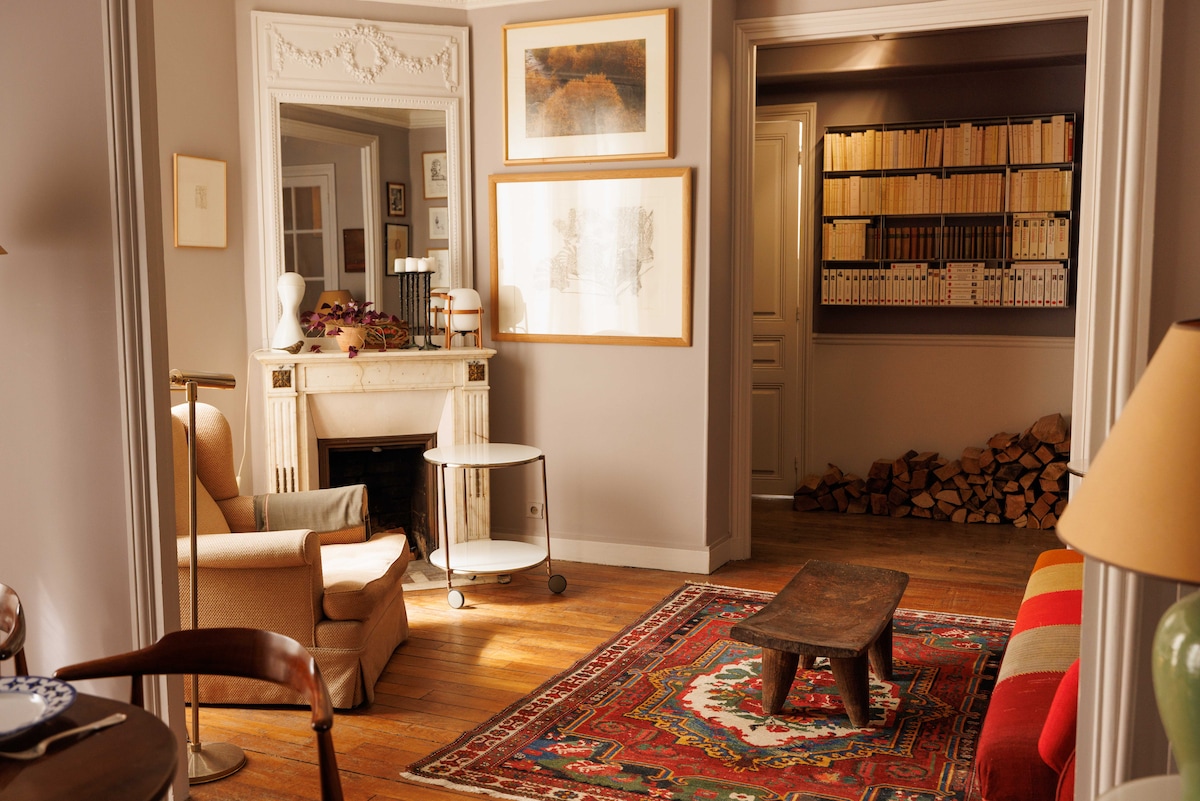 A warm corridor is highlighted by a mix of modern and classic decor. A cozy armchair sits near a minimalist side table and a decorative fireplace. Artworks adorn the walls, and a curated bookshelf is visible, along with a stack of neatly arranged firewood.