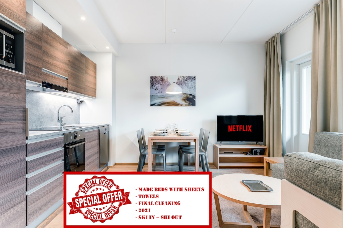 The open-plan living area features a modern kitchen equipped with stainless steel appliances, a dining table for four, and a comfortable lounge space. A large window allows natural light to brighten the room, while a smart TV is positioned adjacent to the seating area.