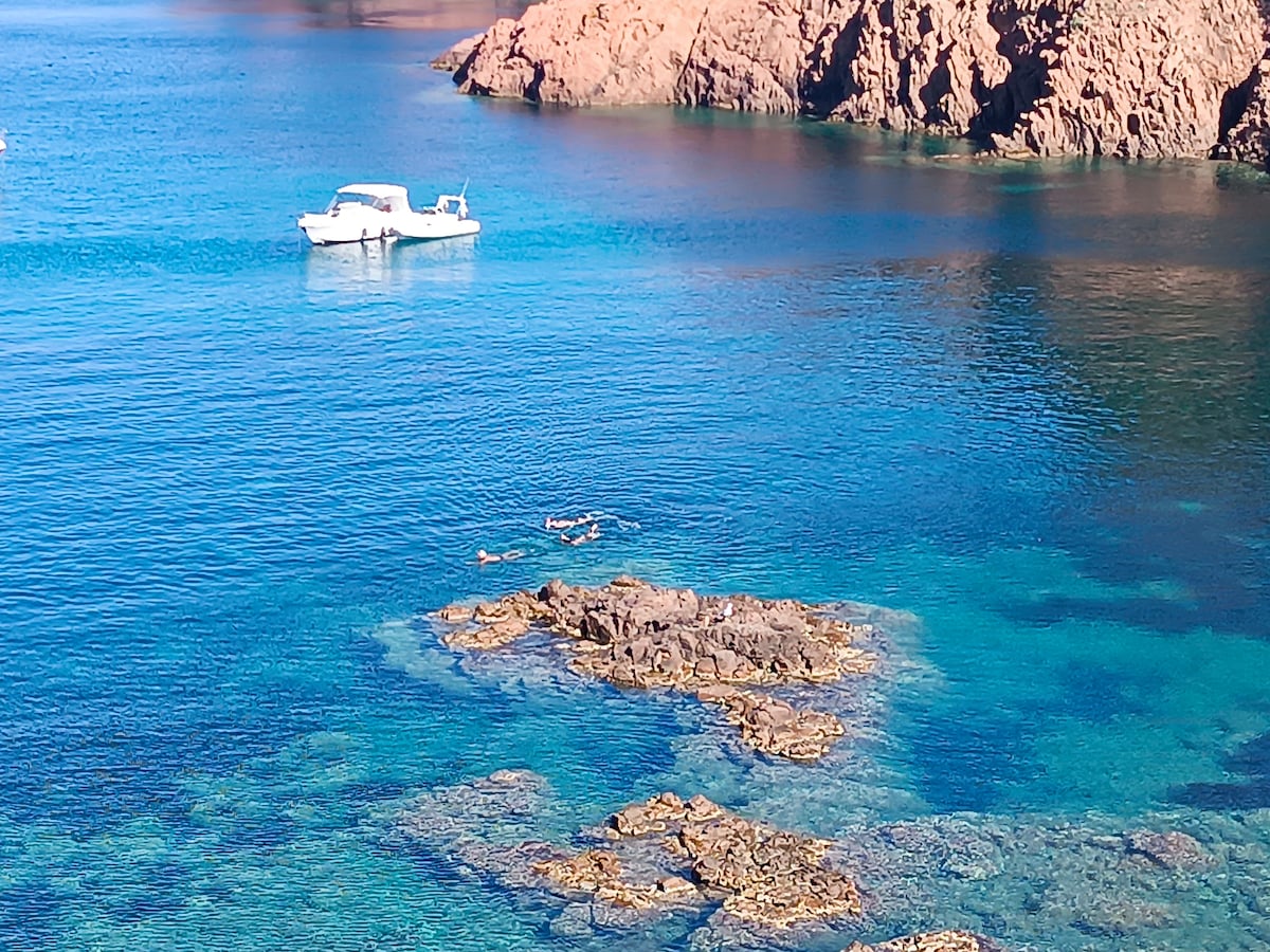 Clear blue waters are visible, with a boat anchored nearby. Two individuals are seen swimming close to rocky formations beneath the surface. The surrounding cliffs create a serene backdrop, enhancing the coastal tranquility of the scene.
