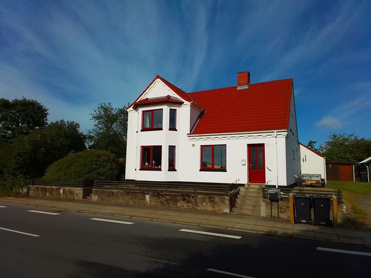 A two-story white house is depicted, featuring a bright red roof and large windows framed in red. Lush greenery surrounds the property, which is bordered by a low stone wall. Trash bins are located at the front, and the setting is bright under a blue sky.