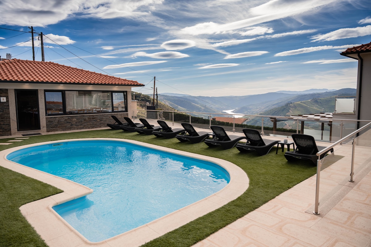 A rectangular pool is surrounded by artificial grass and features several black loungers arranged in a row. The area offers expansive views of a river and mountains under a partly cloudy sky, with a modern building visible in the background.