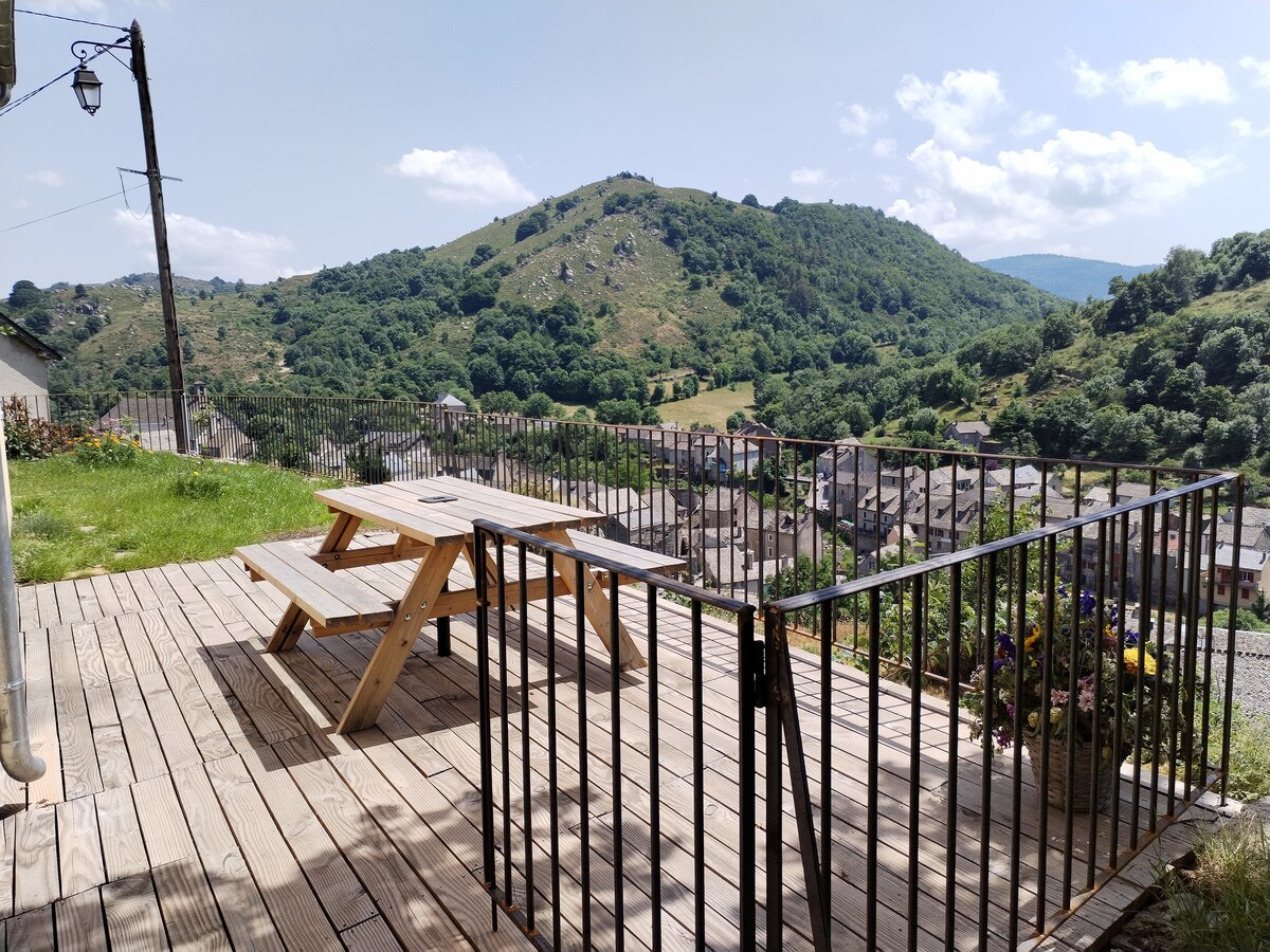 A spacious wooden terrace is enclosed by a black railing, offering a panoramic view of the surrounding hills and valleys. Sunlight illuminates the area, highlighting a wooden picnic table surrounded by greenery, with colorful flowers visible in the planters nearby.