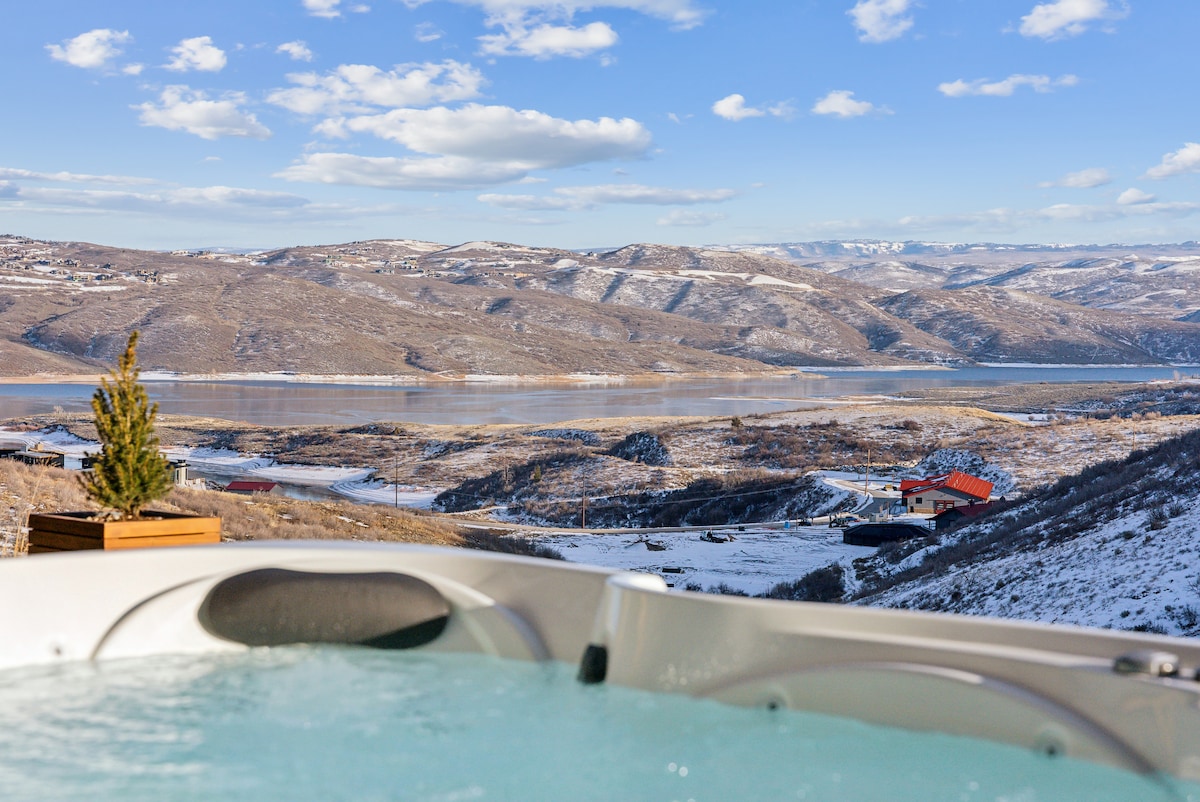 A private hot tub is visible in the foreground, with steam rising against a backdrop of rolling snow-covered hills and a serene lake. Houses are seen dotting the landscape, and a clear blue sky with scattered clouds enhances the peaceful mountain view.