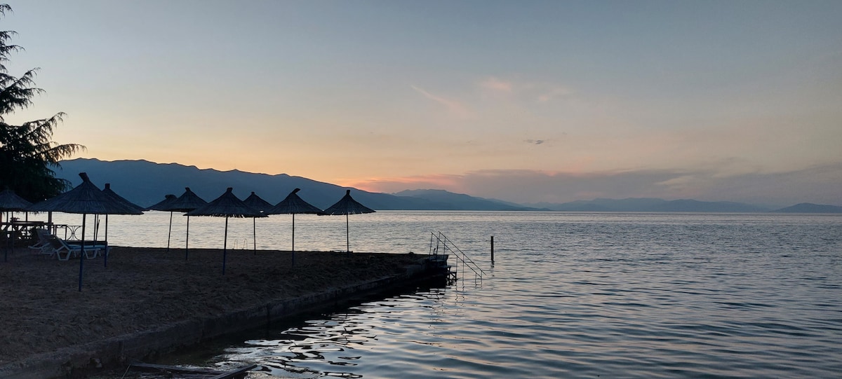 A serene lakeside view is presented, featuring several thatched umbrellas along a sandy shore. The calm water reflects the soft hues of the sunset, with distant mountains visible on the horizon. A gentle slope leads into the water, alongside a sturdy staircase.