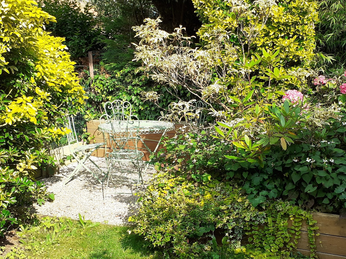 A serene garden area features a small gravel section surrounded by lush greenery. A delicate wrought iron table and four matching chairs are positioned amid blooming plants, creating an inviting spot for outdoor relaxation. Sunlight filters through the surrounding foliage.