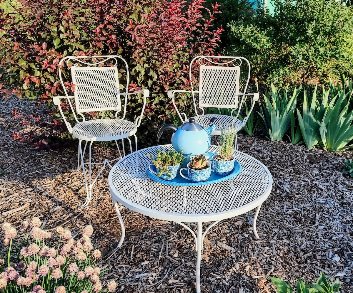 A peaceful garden area features a round metal table topped with a blue teapot and potted succulents. Two white, metal chairs with intricate patterns are positioned on either side of the table. Lush greenery and flowering plants surround the setting, enhancing a serene outdoor environment.
