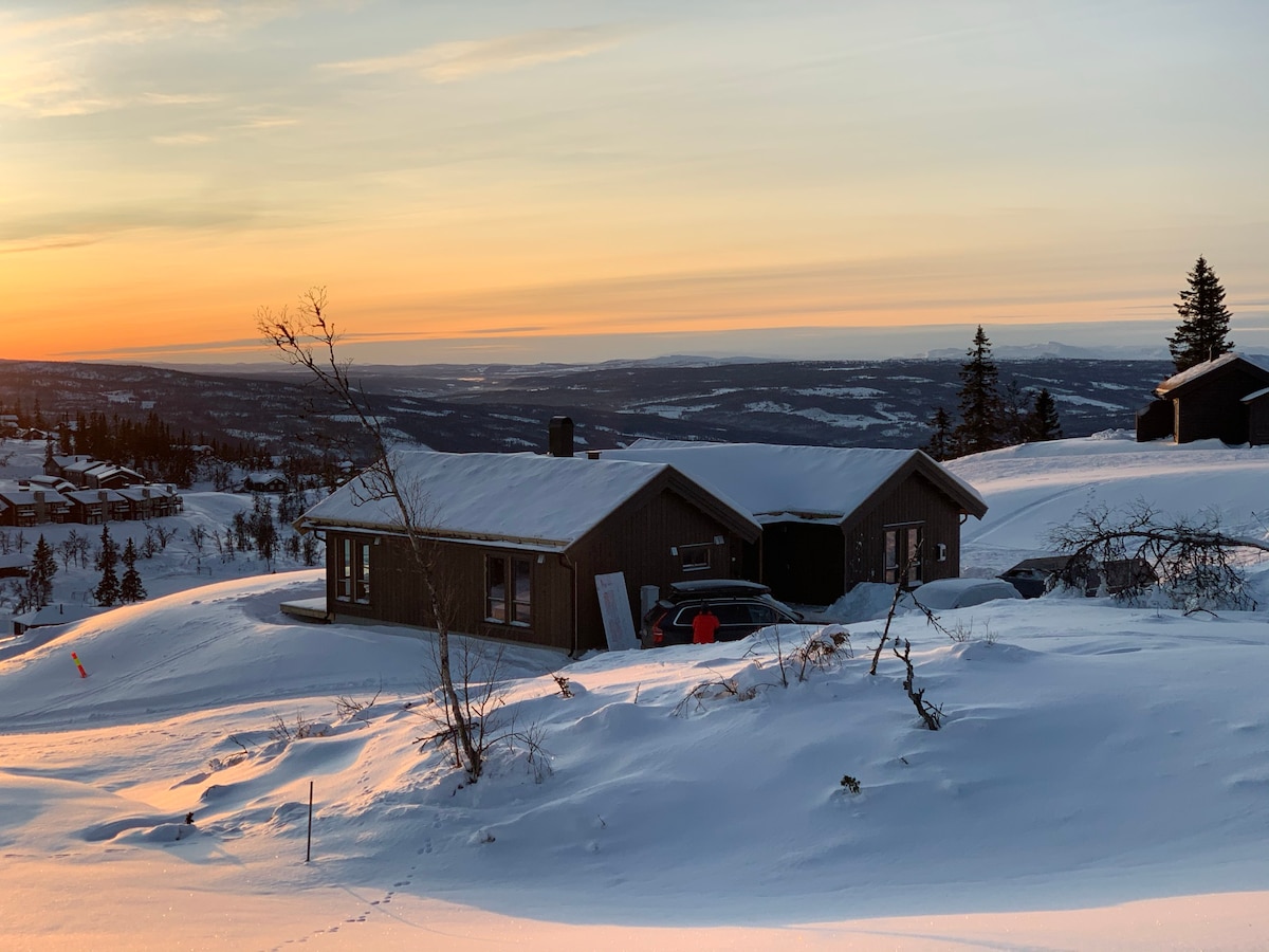 The cabin is nestled in a snowy landscape, surrounded by gentle slopes and sparse trees. A warm sunset casts an orange glow over the horizon, highlighting the peaceful winter scenery. Nearby vehicles are parked, and the distant mountains are faintly visible.