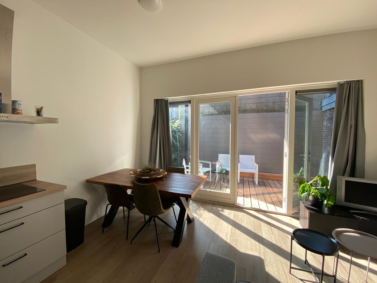 A dining area is positioned near a large glass door that opens to a private outdoor space. A wooden table with stylish chairs is present, accompanied by a small potted plant. Natural light brightens the room, enhancing the functional kitchen space adjacent to it.