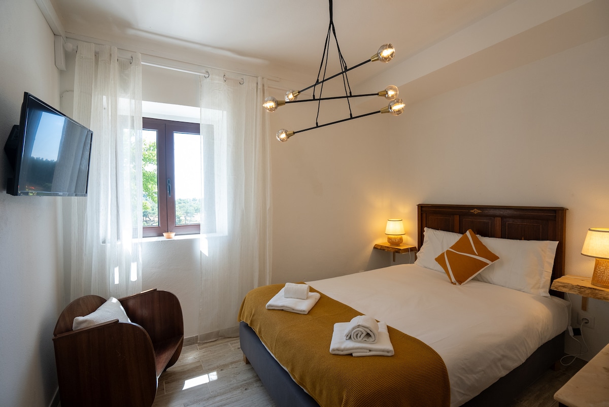 A well-lit bedroom features a spacious bed with white linens and a mustard-colored throw pillow. A wooden headboard is complemented by bedside tables with lamps. Natural light filters through sheer curtains, illuminating a comfortable chair and a small television mounted on the wall.