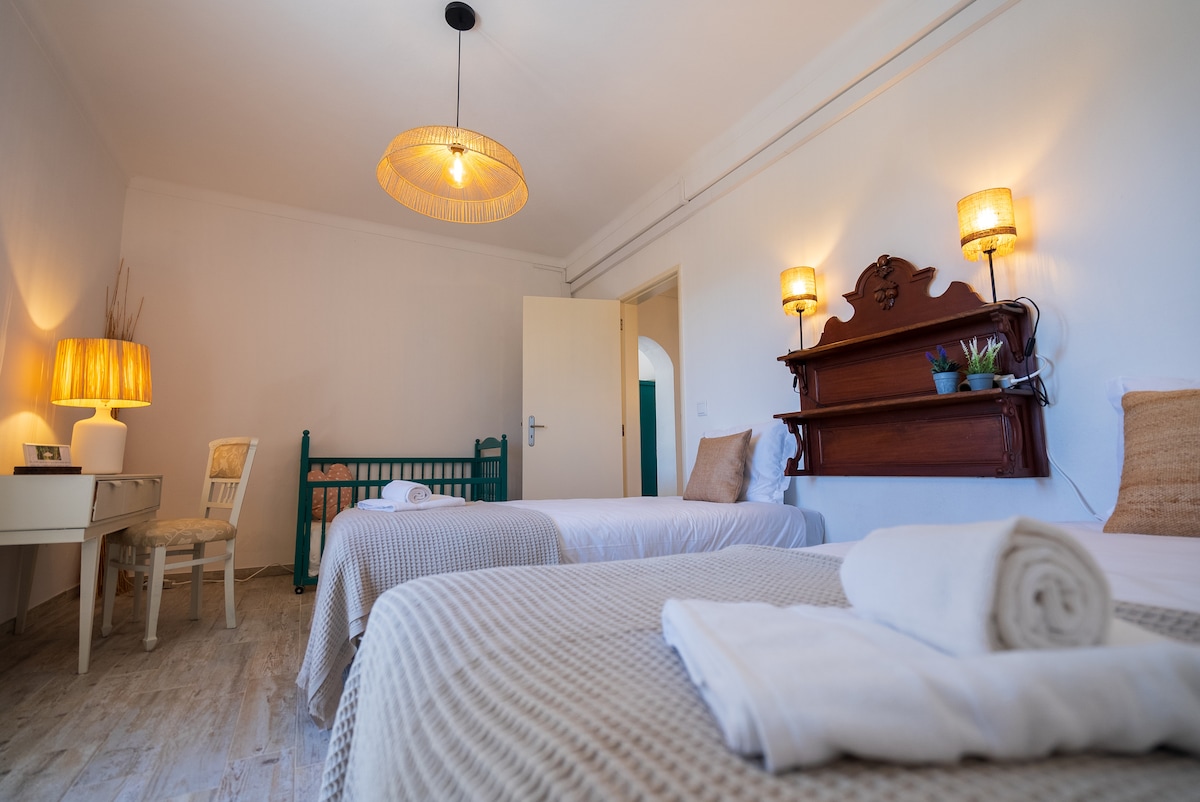A guest room features two individual beds neatly made with white linens. A small desk with a lamp sits beside one bed, while a decorative shelf holds potted plants. Soft lighting is provided by two wall sconces, and a cozy crib is positioned in the corner.